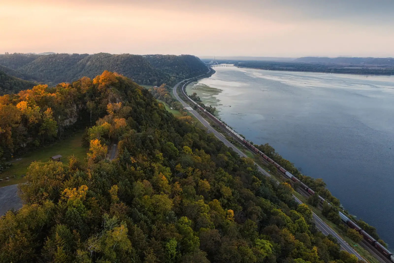 Wooded bluffs in autumn colour above a wide bend of the Mississippi River, with a highway and freight rail line running along the bank