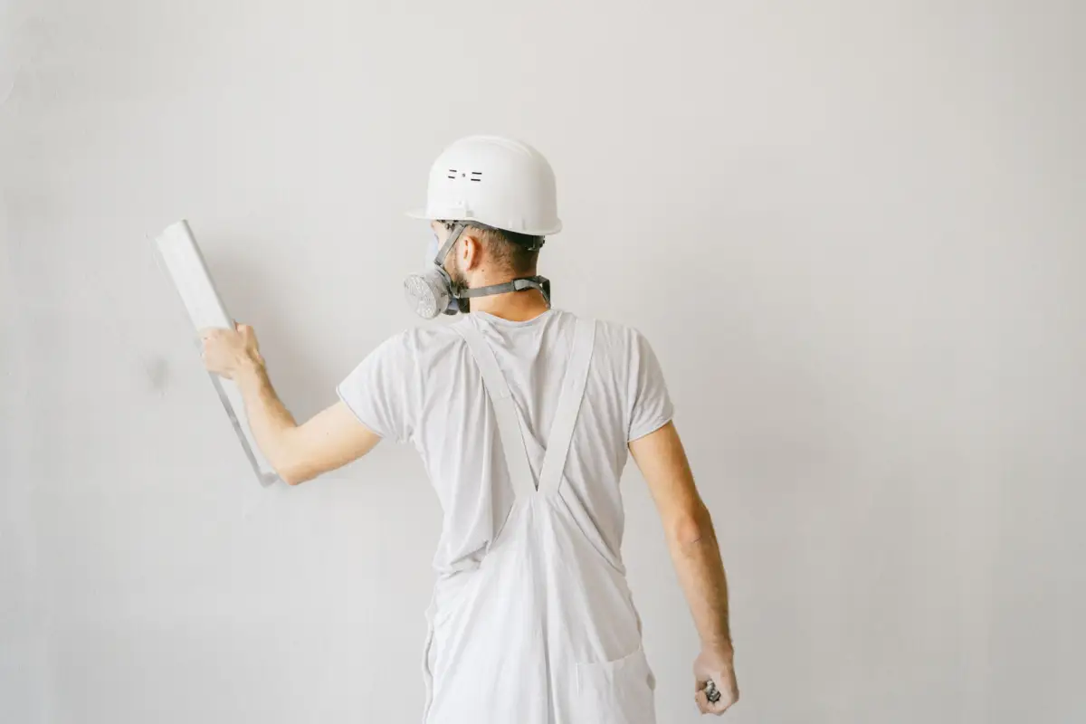 Drywall finisher in a hard hat and respirator skimming joint compound across a wall with a wide blade